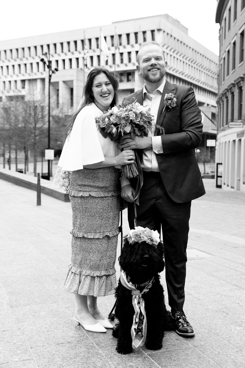 Black and white photo of smiling newlyweds holding bouquet with their dog wearing floral crown outside Boston City Hall.