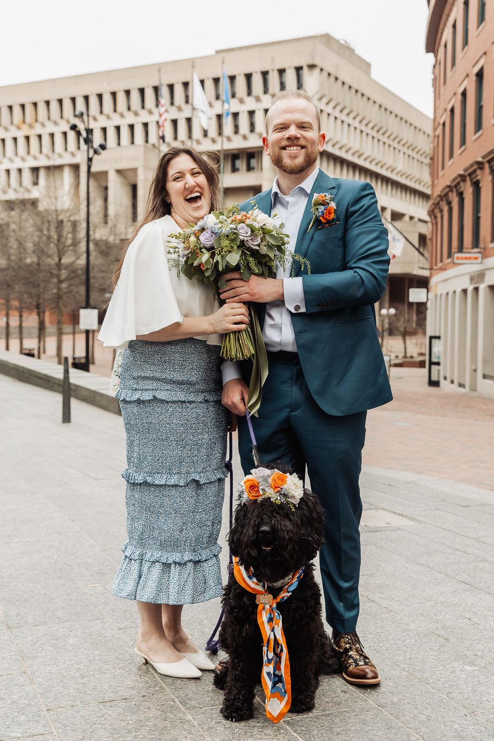 Happy couple in wedding attire poses with black dog outside Boston City Hall, bride holds floral bouquet.