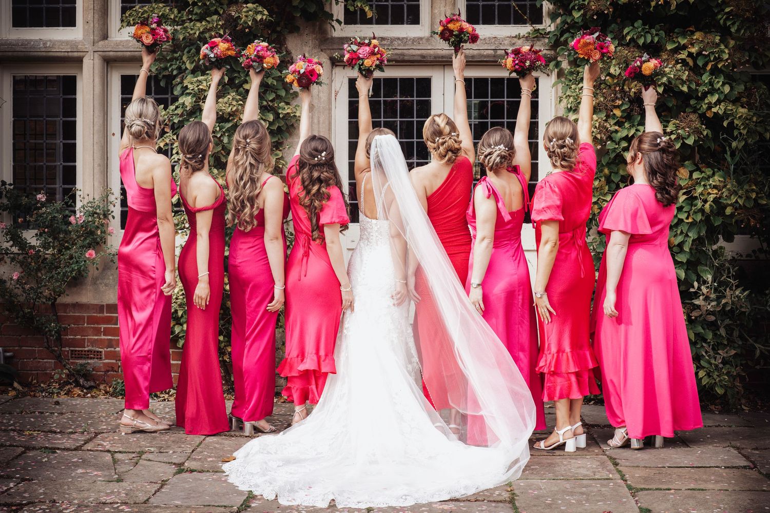 Bride in white gown and veil poses with bridesmaids in red and pink dresses holding bouquets aloft outdoors.