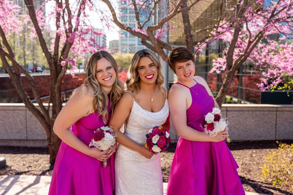 Bride in white gown flanked by two bridesmaids in fuchsia dresses holding bouquets under pink flowering trees  in downtown Omaha, NE