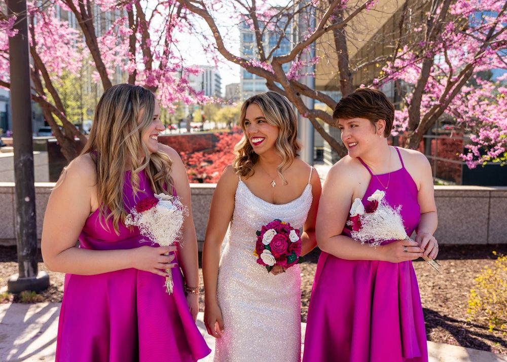 Bride in white gown smiles with two bridesmaids in fuchsia dresses holding bouquets under spring blooming pink trees in downtown Omaha, NE