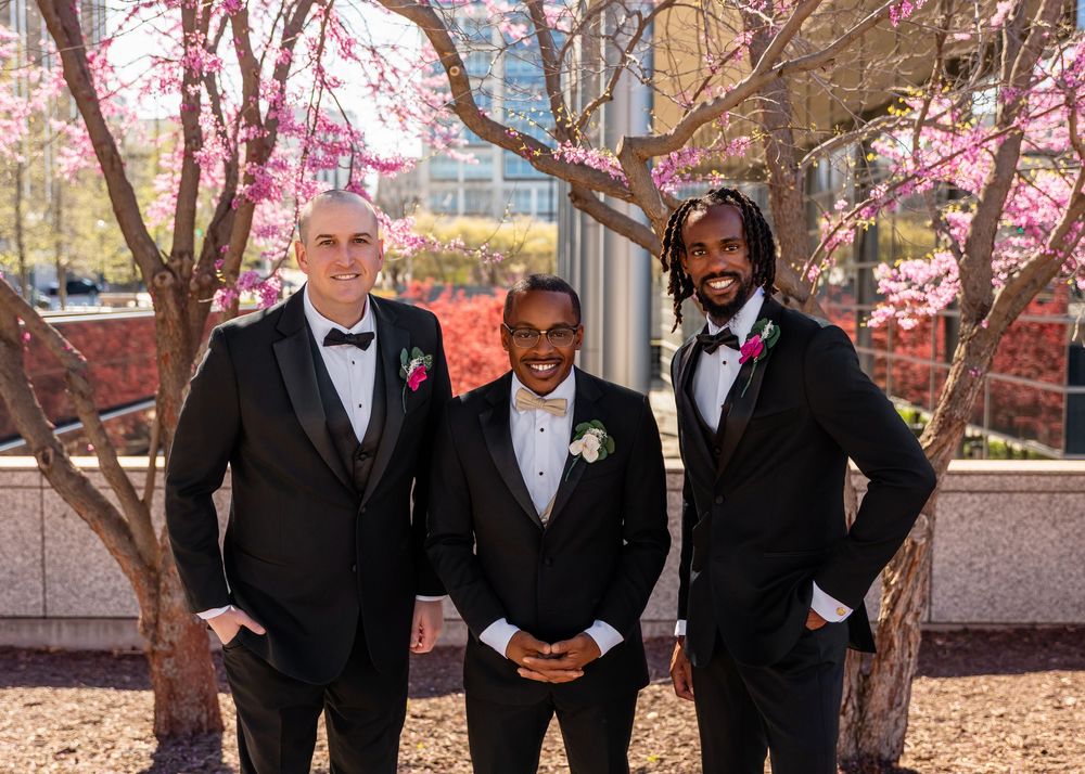 Three groomsmen in black tuxedos with pink boutonnieres pose outdoors under blooming pink cherry blossom trees  in downtown Omaha, NE.