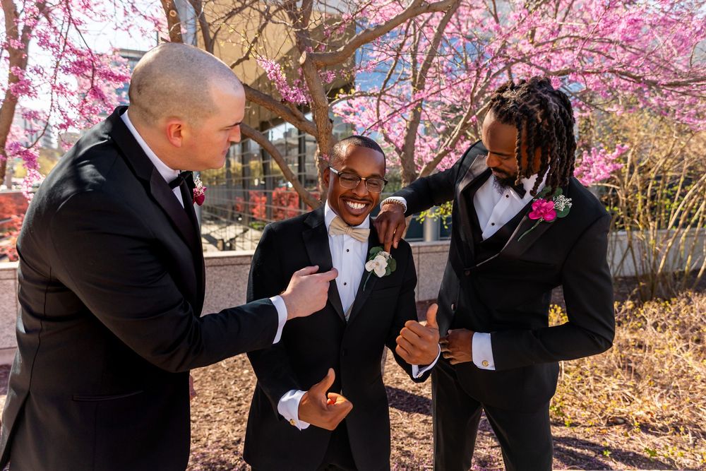 Three groomsmen in black tuxedos laugh and celebrate together under blooming pink cherry blossom trees outdoors  in downtown Omaha, NE.