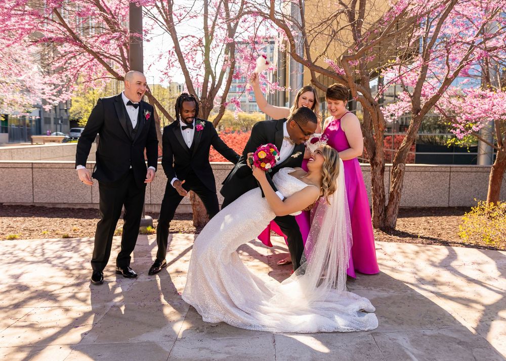 Wedding party celebrates as bride and groom share a dip kiss under blooming pink cherry blossom trees  in downtown Omaha, NE