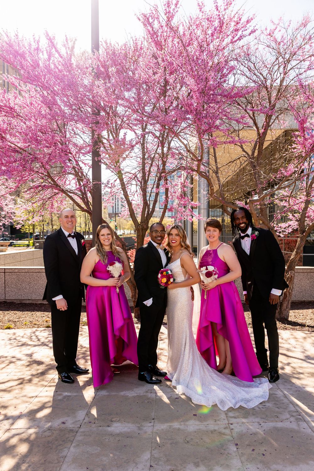 Wedding party poses before pink blooming trees, bride in white gown with bridesmaids in fuchsia dresses and groomsmen in tuxedos  in Omaha
