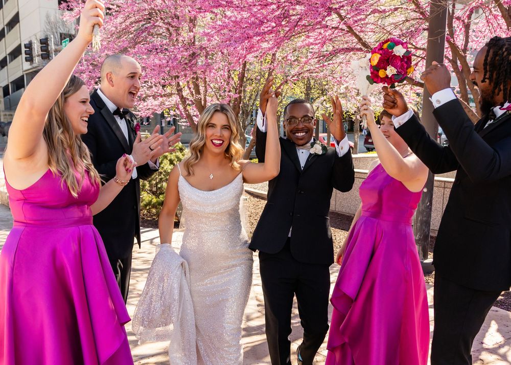 Joyful wedding party celebrating outdoors with pink cherry blossoms, bride in white gown, groom in tux, bridesmaids in fuchsia.