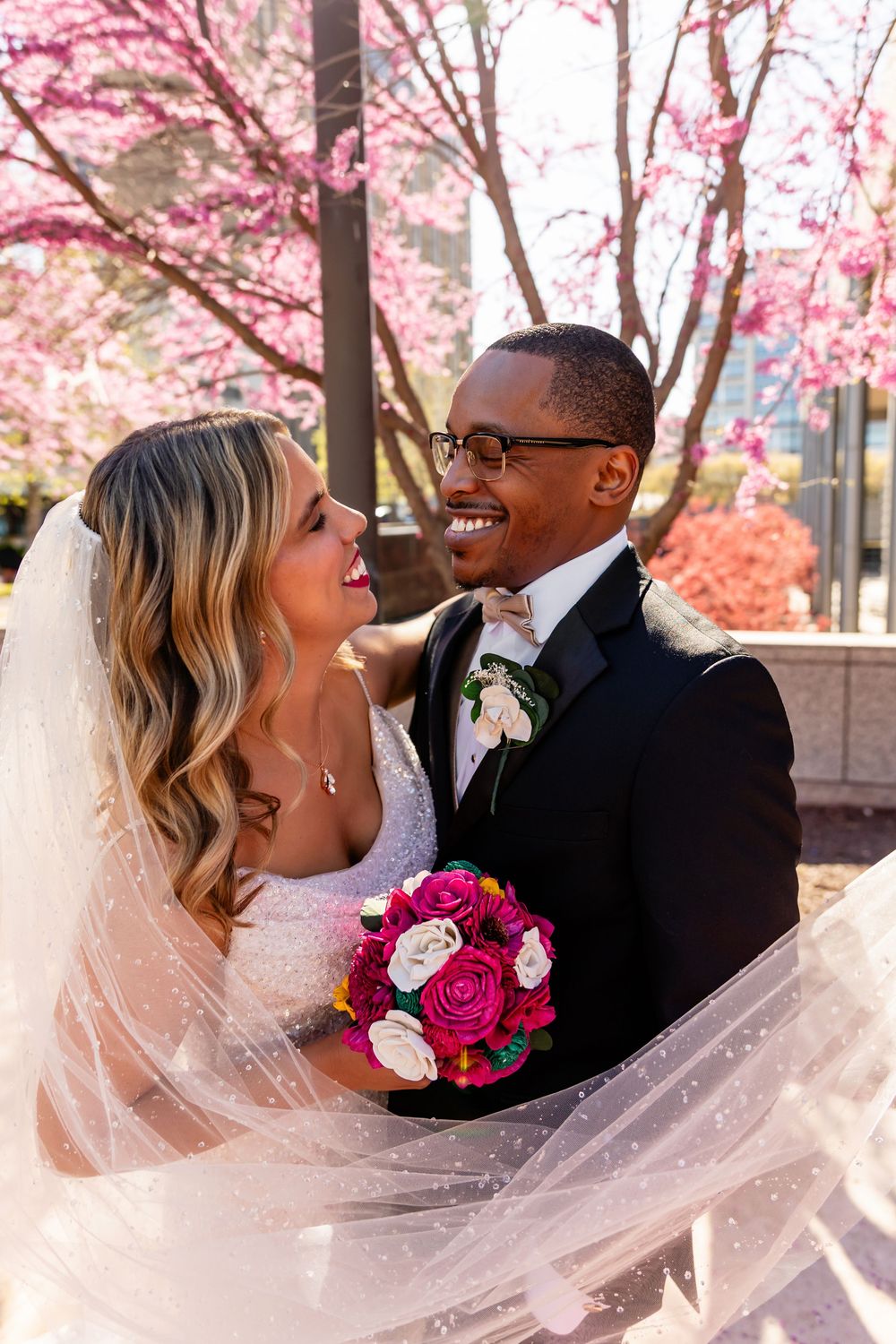 Smiling bride and groom embrace outdoors with pink cherry blossoms, bride holding vibrant fuchsia and white bouquet  in downtown