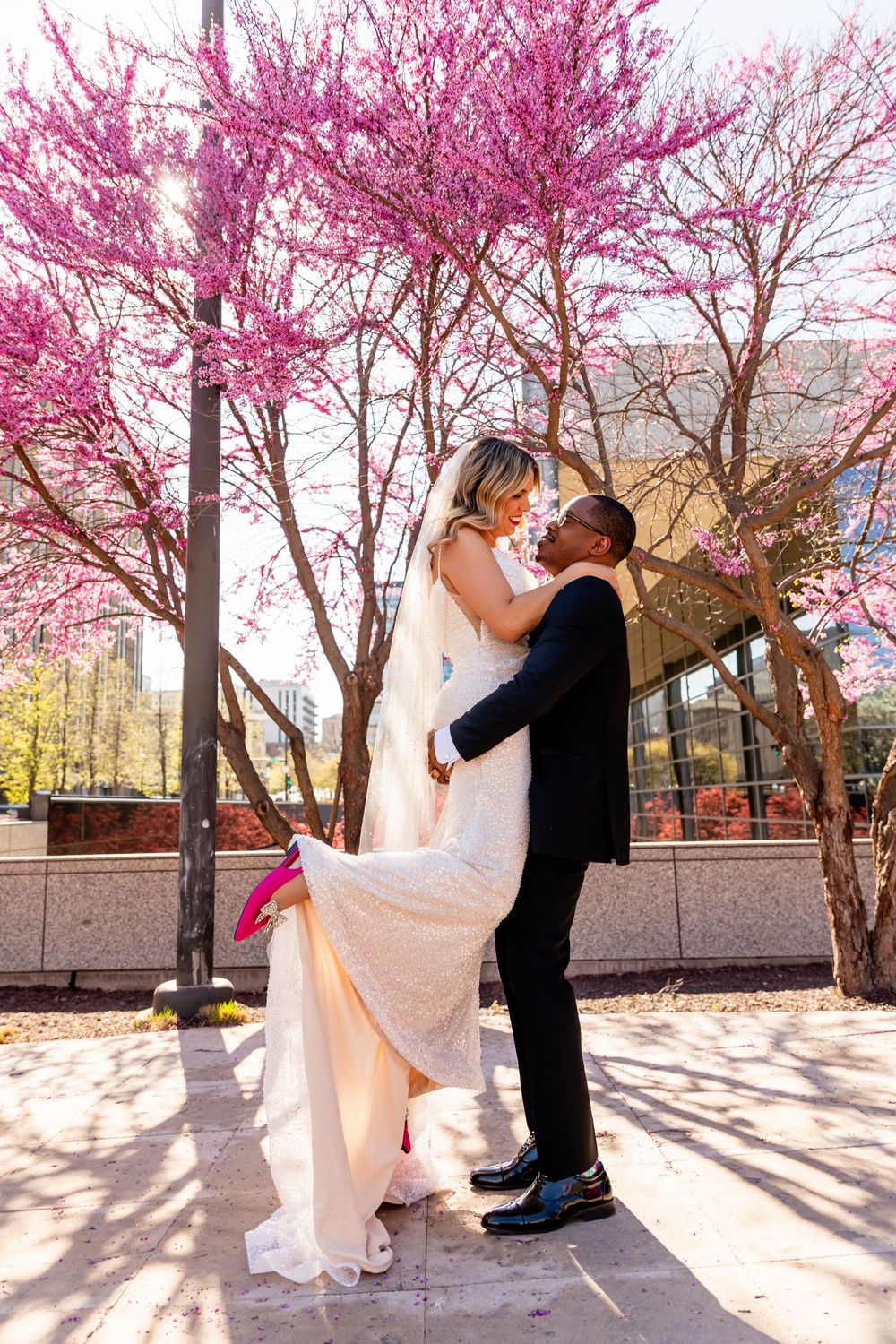 Bride and groom share a romantic lift kiss beneath blooming pink cherry blossom trees on their spring wedding day in downtown Omaha, NE