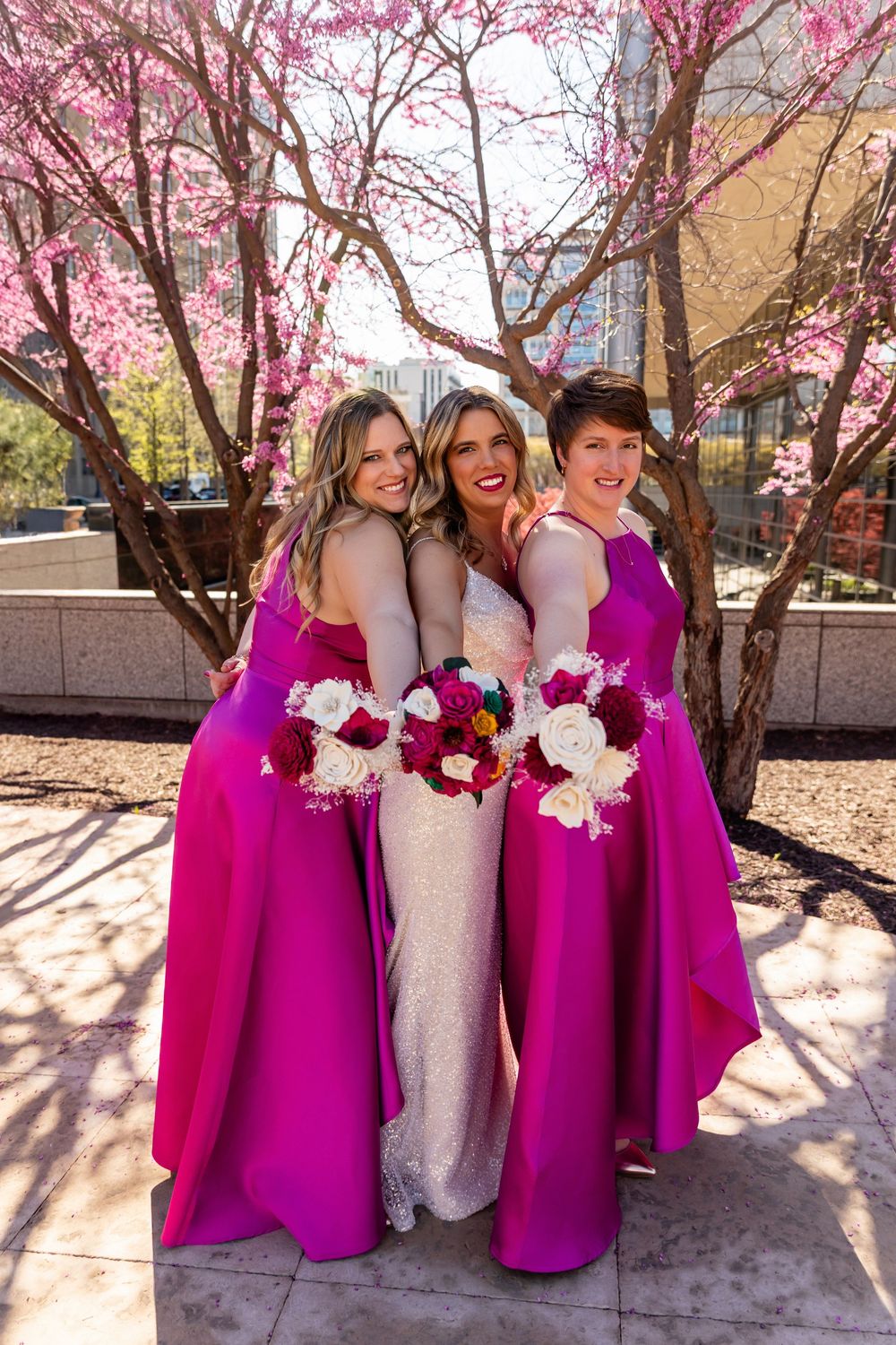 Bride and bridesmaids in magenta gowns pose from behind showing bouquets under flowering cherry blossom trees  in downtown Omaha, NE