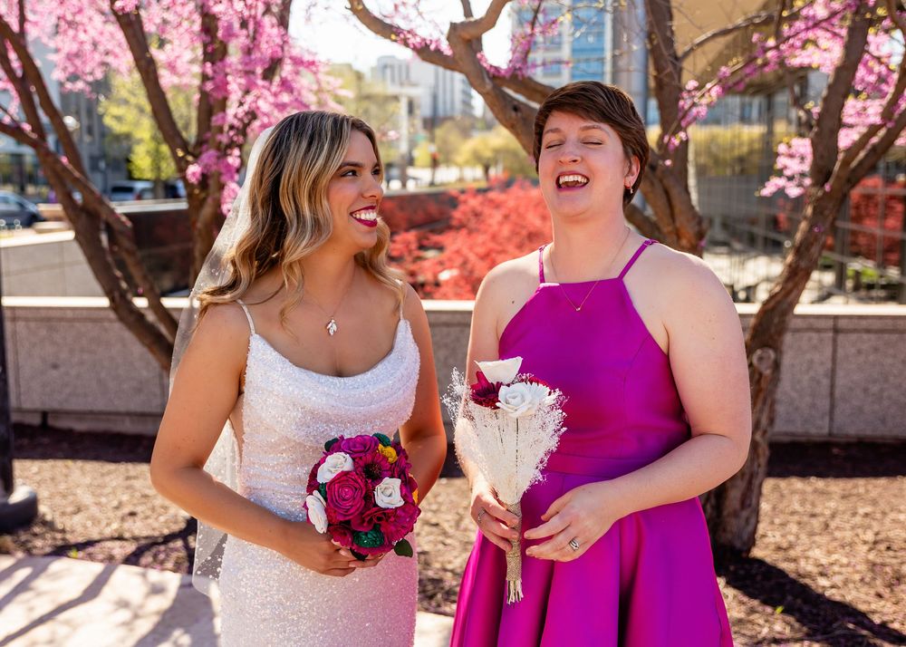 Two women laughing outdoors in wedding attire, one in white gown and one in magenta dress, holding bouquets under pink blooming trees
