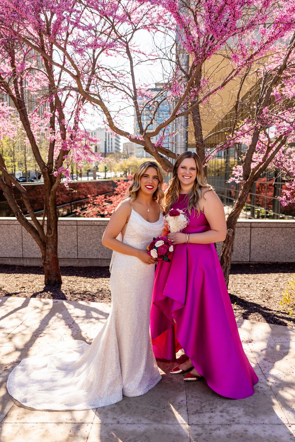 Bride in white lace gown poses with bridesmaid in fuchsia dress under vibrant pink blooming trees outdoors  in downtown Omaha, NE
