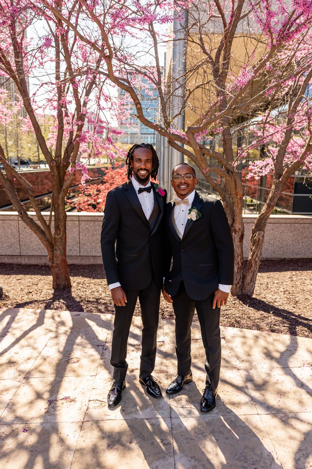Two grooms in black tuxedos smiling outdoors with pink cherry blossom trees in spring wedding photo  in downtown Omaha, NE