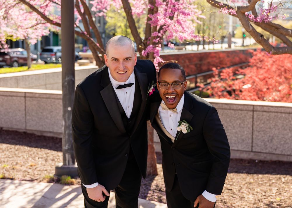 Two men in black tuxedos smiling together outdoors under pink cherry blossom trees at a wedding celebration  in downtown Omaha, NE