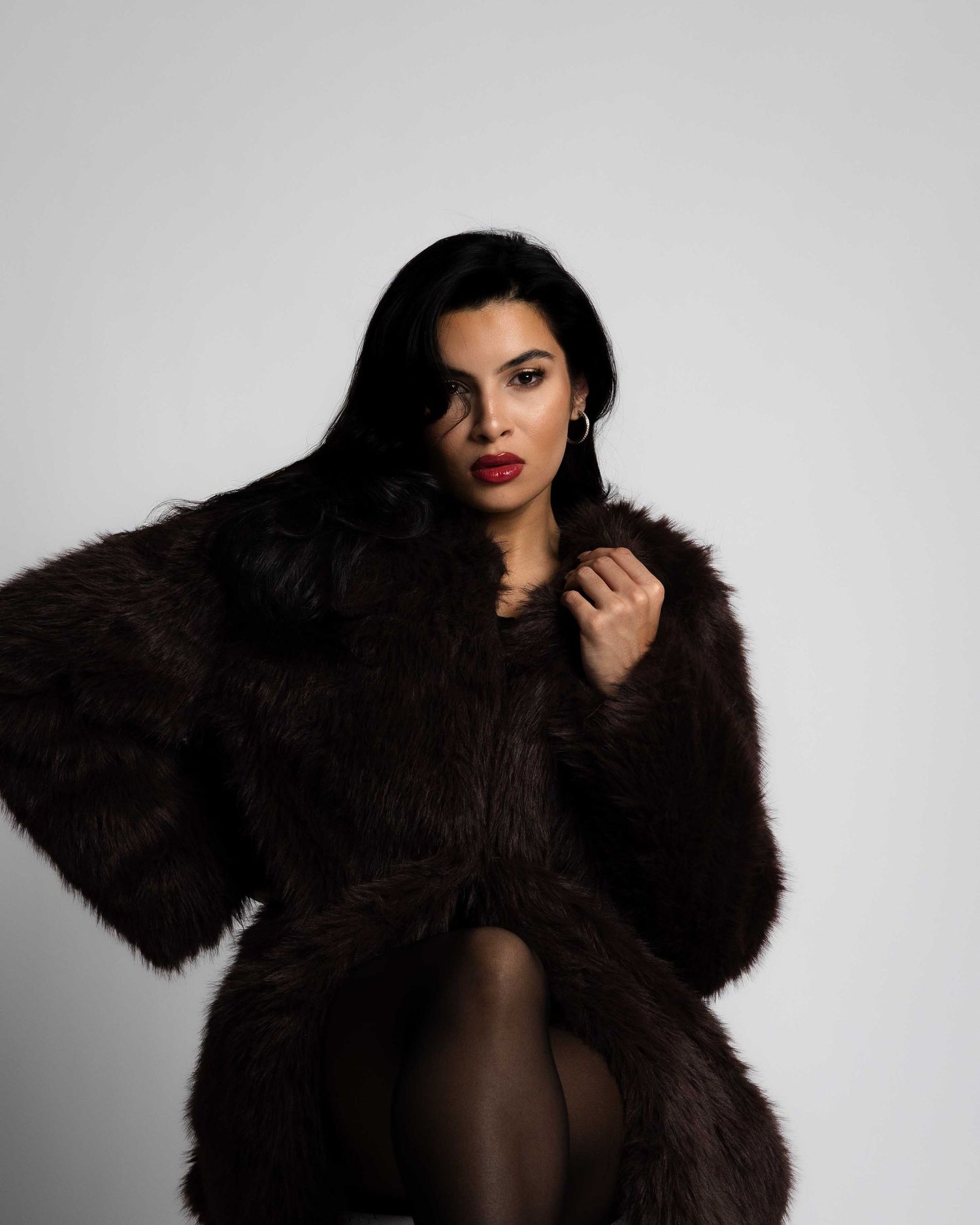 A model in a luxurious dark fur coat poses against a light gray studio background in a fashion editorial series.