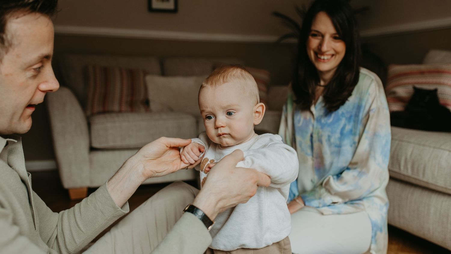 A baby plays and laughs with parents on a cozy beige couch in a living room setting.