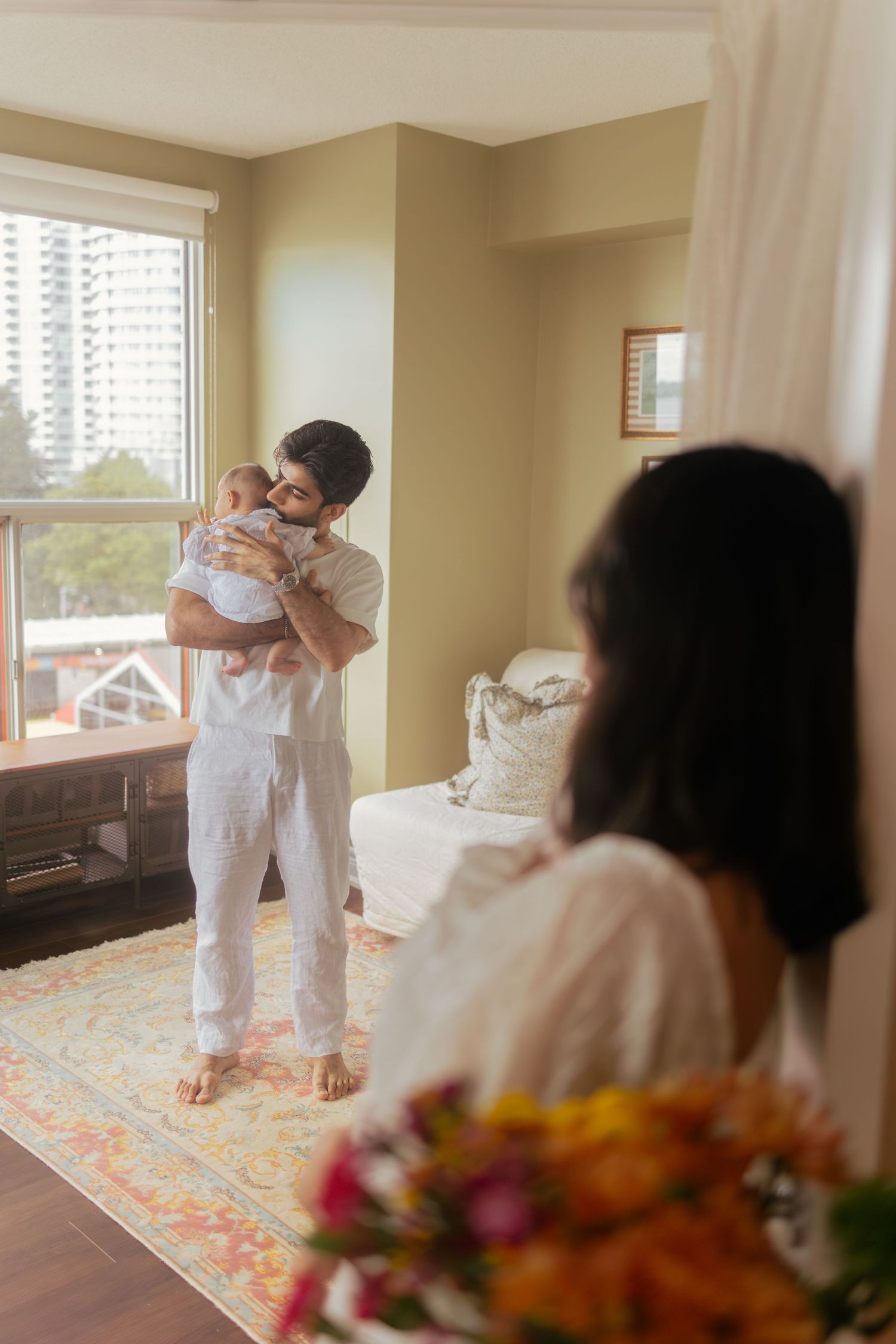 Wedding party helps bride put on white dress in hotel room with floral carpet and window light.