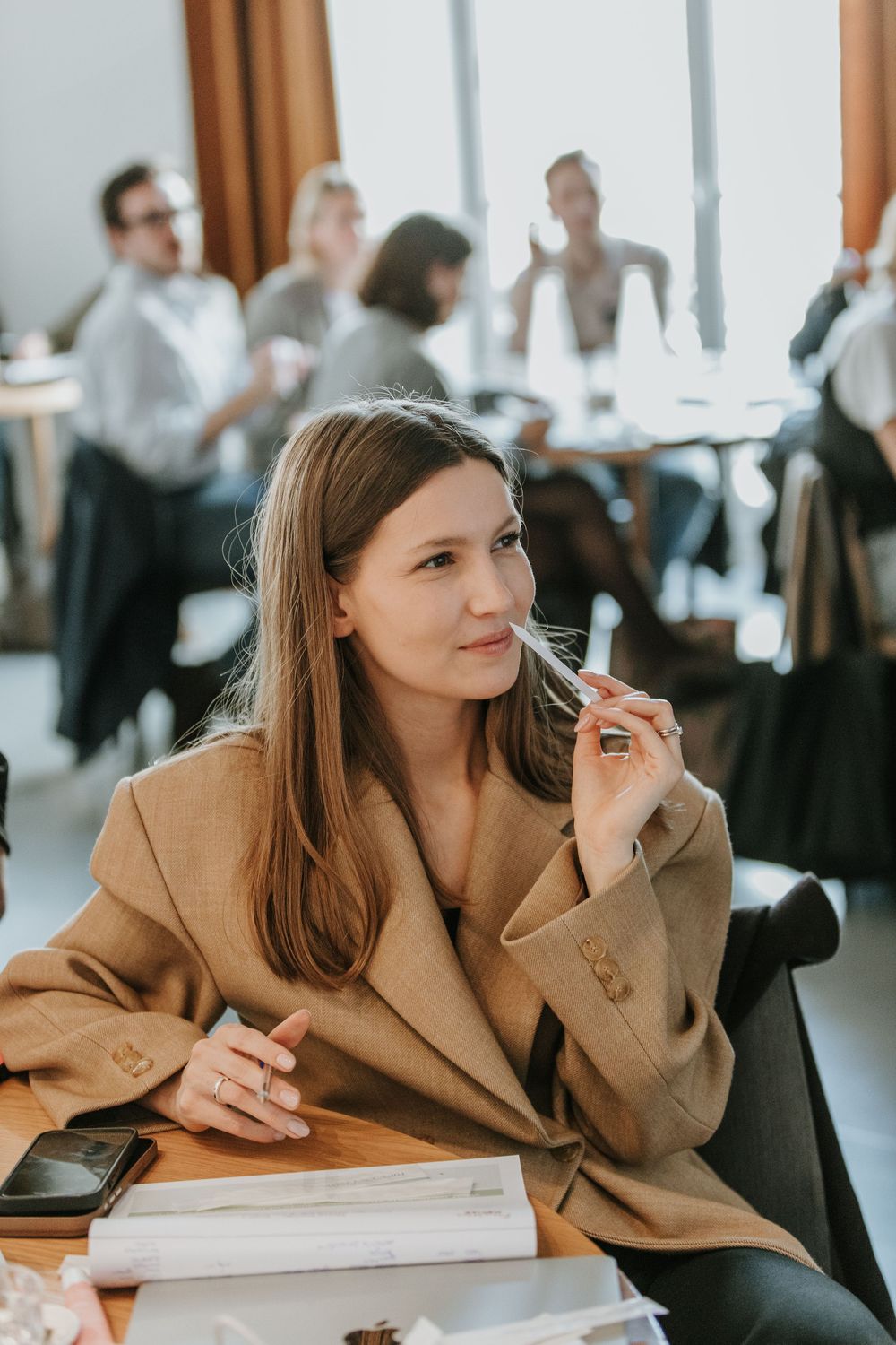 Professional wearing a tan blazer sits thoughtfully at a desk in a bright office setting.