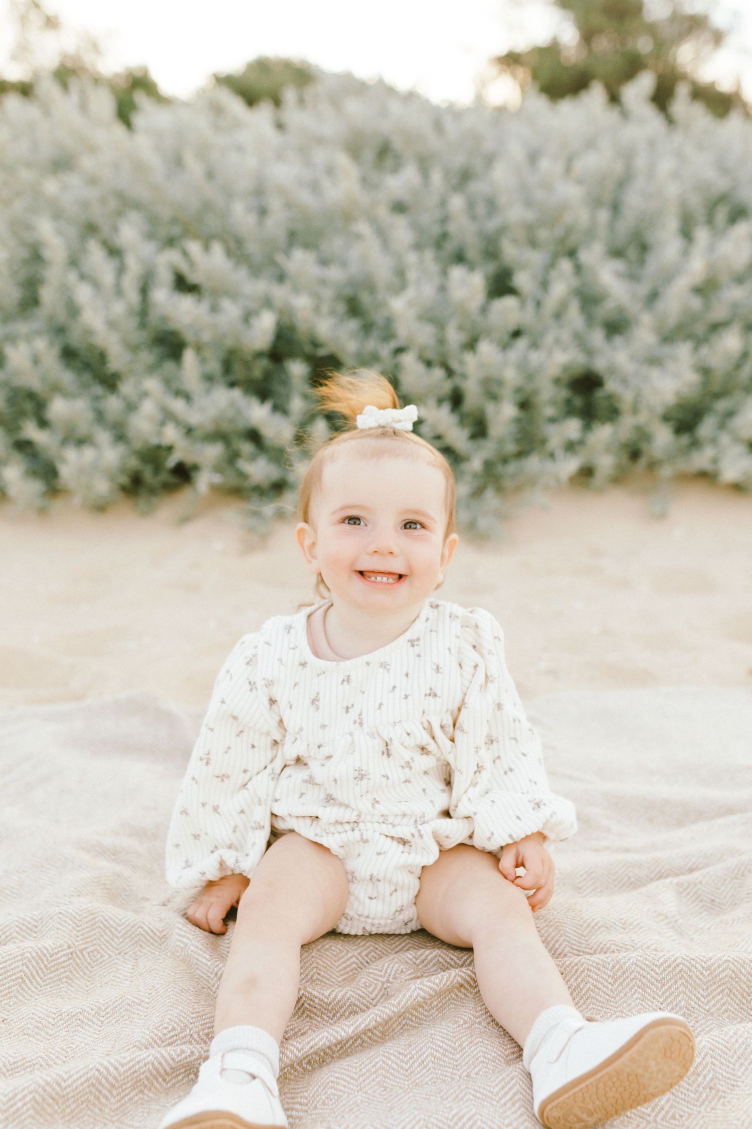 Baby in floral dress crawls and plays on blanket in outdoor setting with white wildflowers in background.