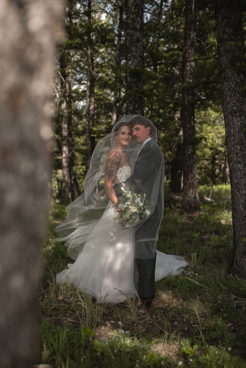 A romantic wedding couple embraces in a sunlit forest setting with a flowing white veil caught in the breeze.
