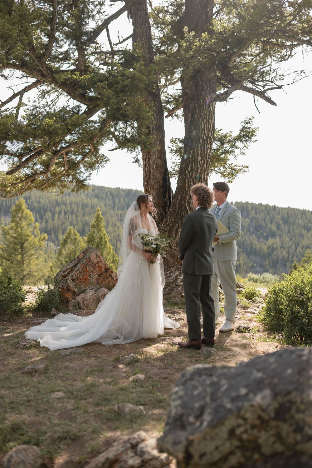 A wedding ceremony takes place on a scenic mountain overlook with tall pine trees and forest views in the background.