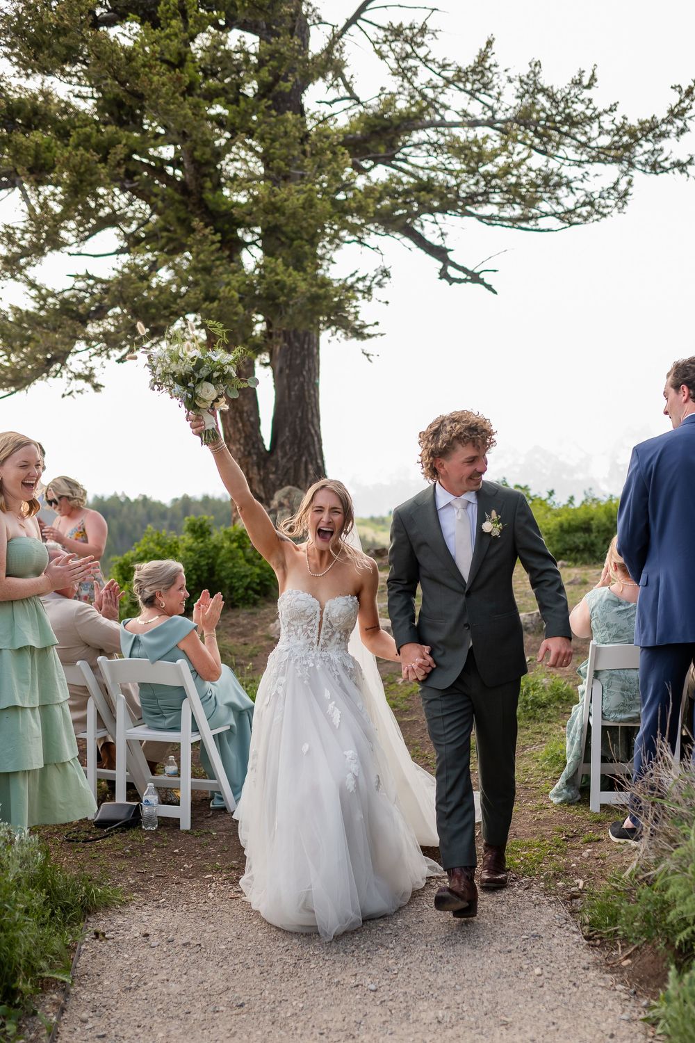 Wedding party celebrates joyfully under a large pine tree during an outdoor ceremony on a scenic hilltop.