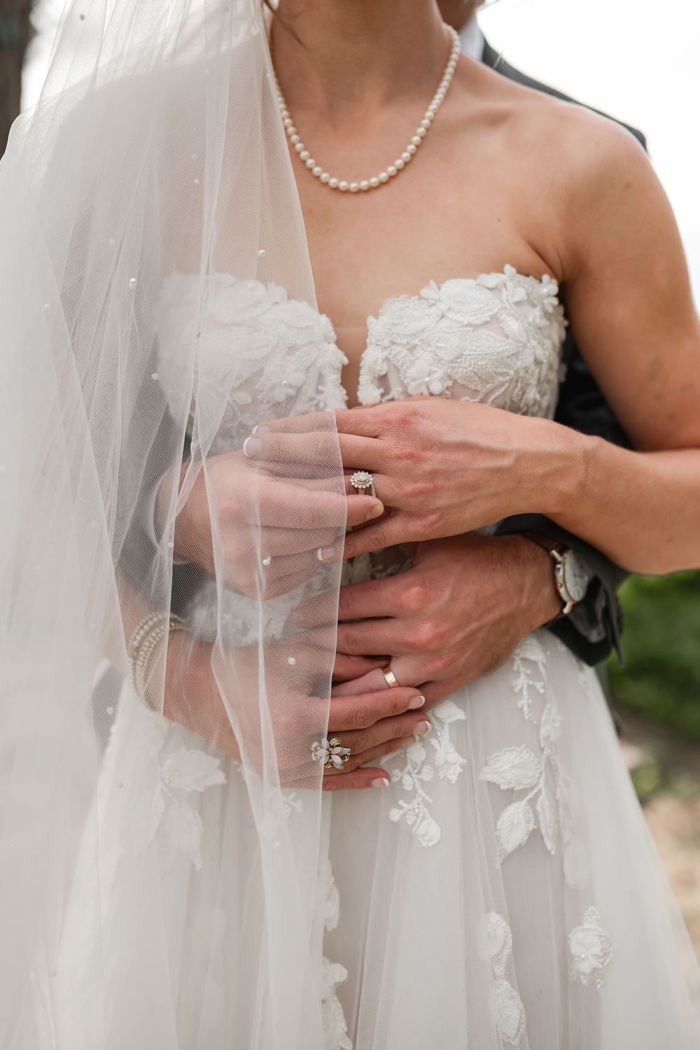 Close-up detail of delicate white lace and tulle wedding dress with pearl necklace and engagement ring.