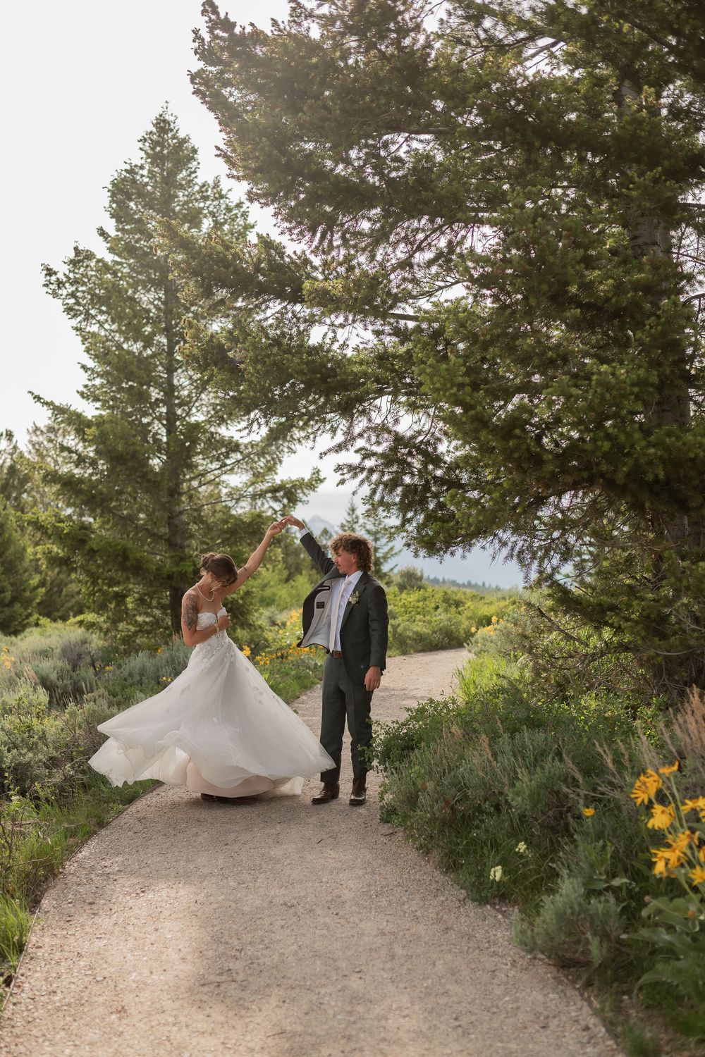 A couple in wedding attire walks along a scenic mountain path surrounded by evergreen trees and yellow wildflowers.