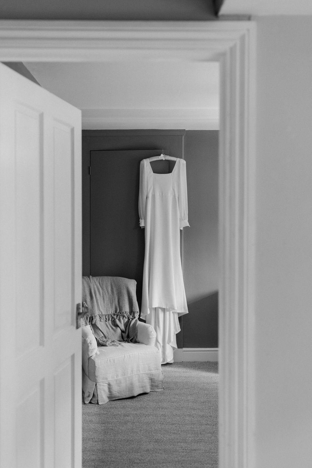Wedding dress hanging in doorway with armchair in monochrome.