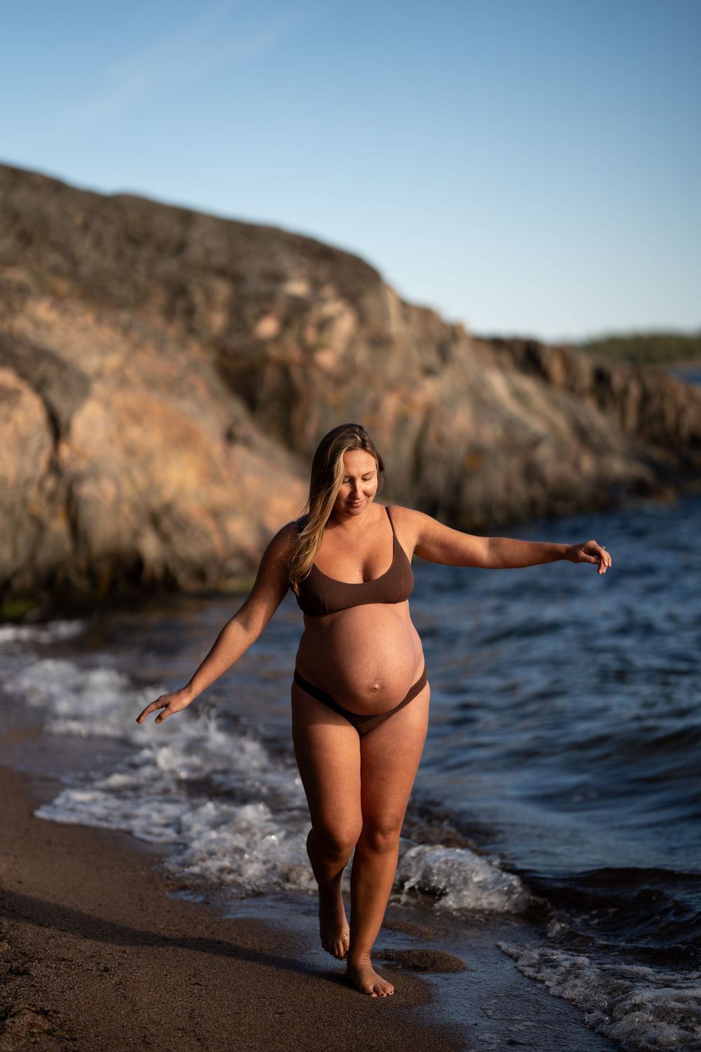 En gravid kvinna går balansgång på en strand. I bakgrunden syns klippor, blå himmel och hav.
