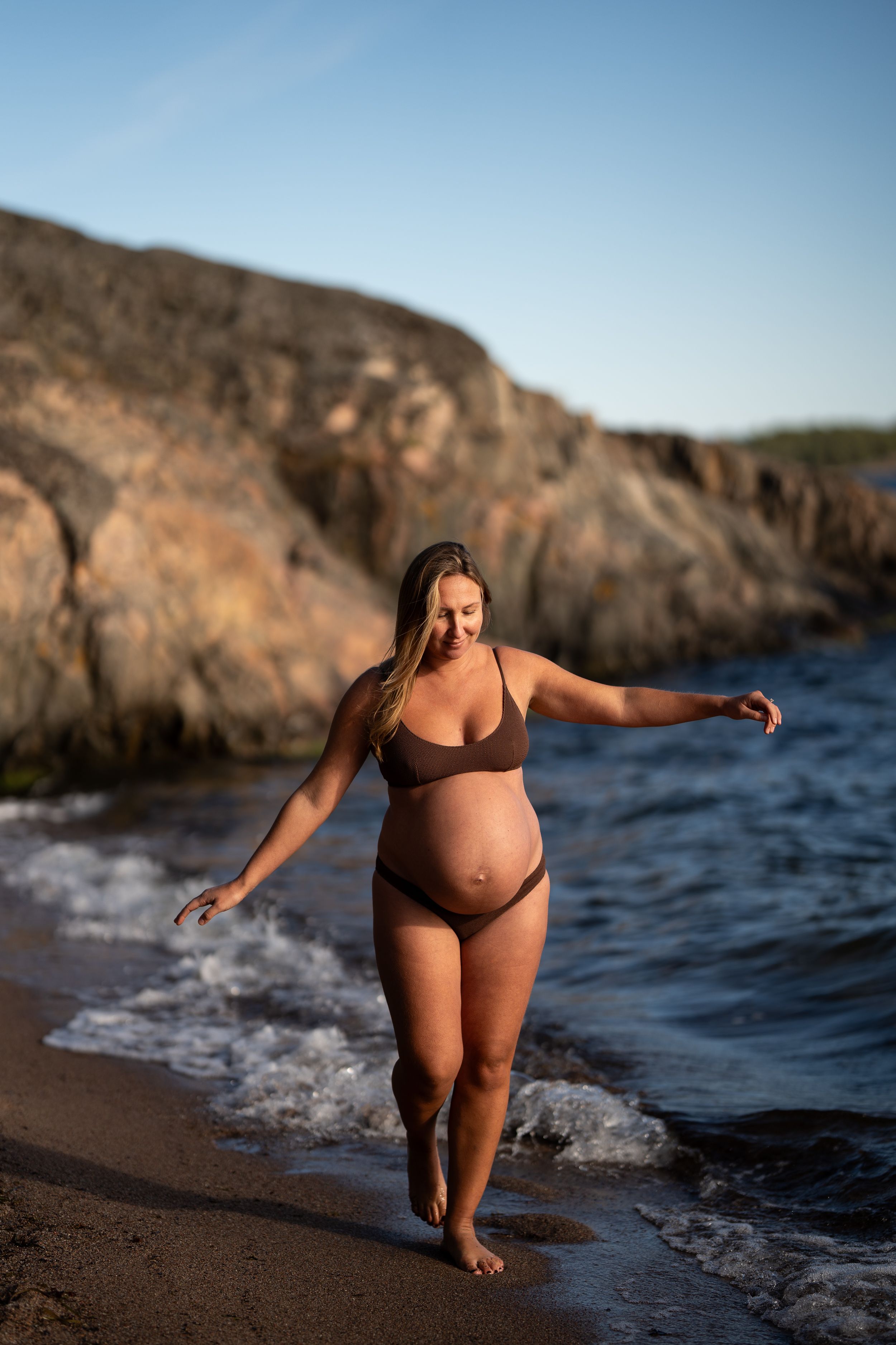 En gravid kvinna går balansgång på en strand. I bakgrunden syns klippor, blå himmel och hav.