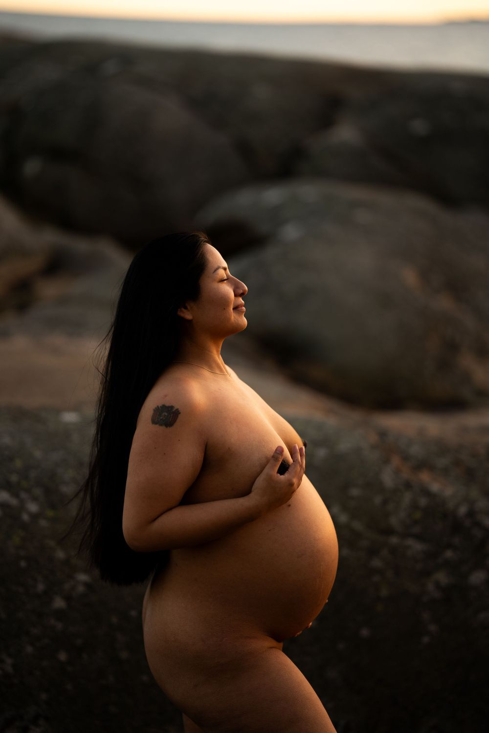 En gravid kvinna står i kvällssolen framför en klippa och håller ena handen på bröstet.