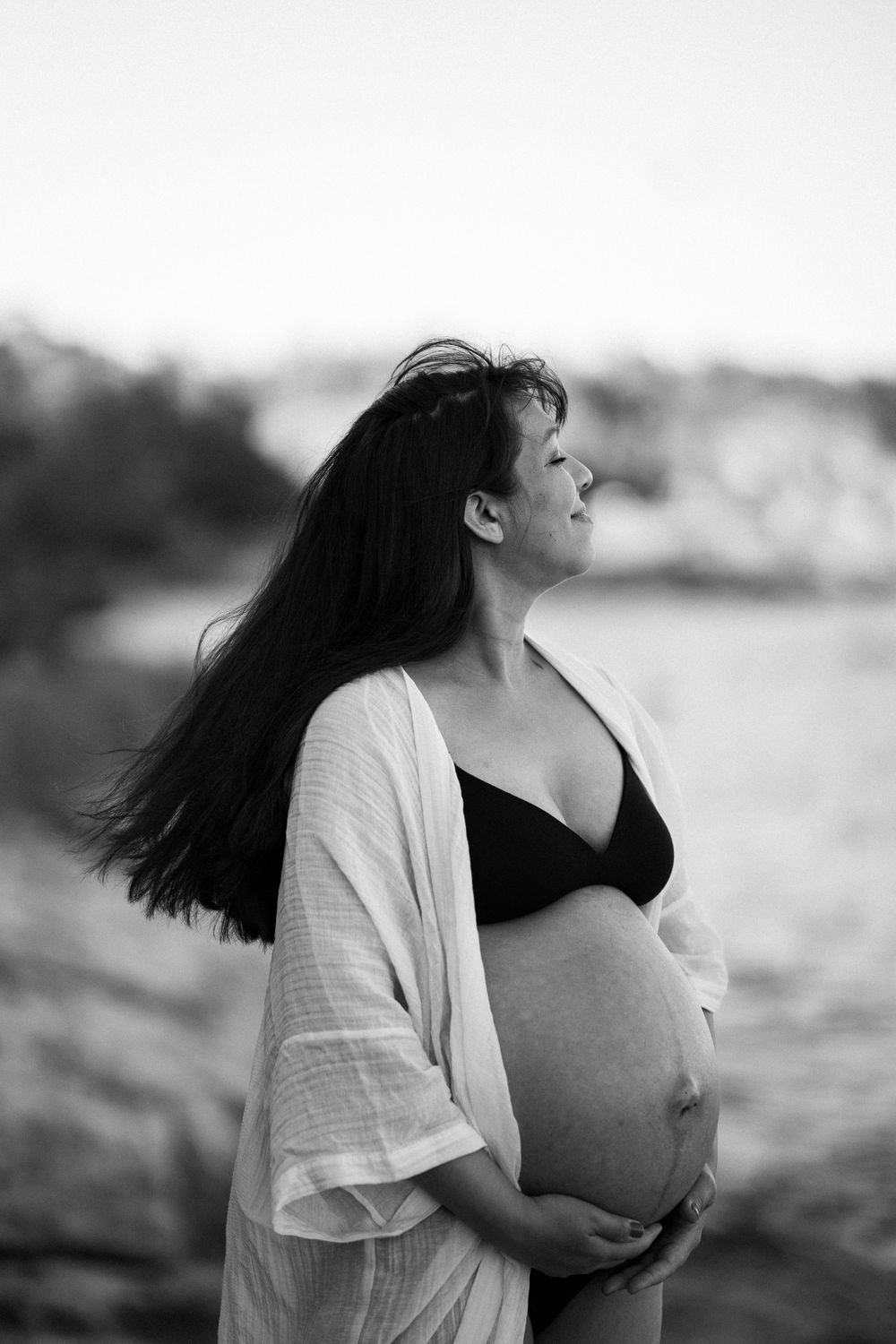 En gravid kvinna med svart topp och vit kimono blundar och håller på magen.