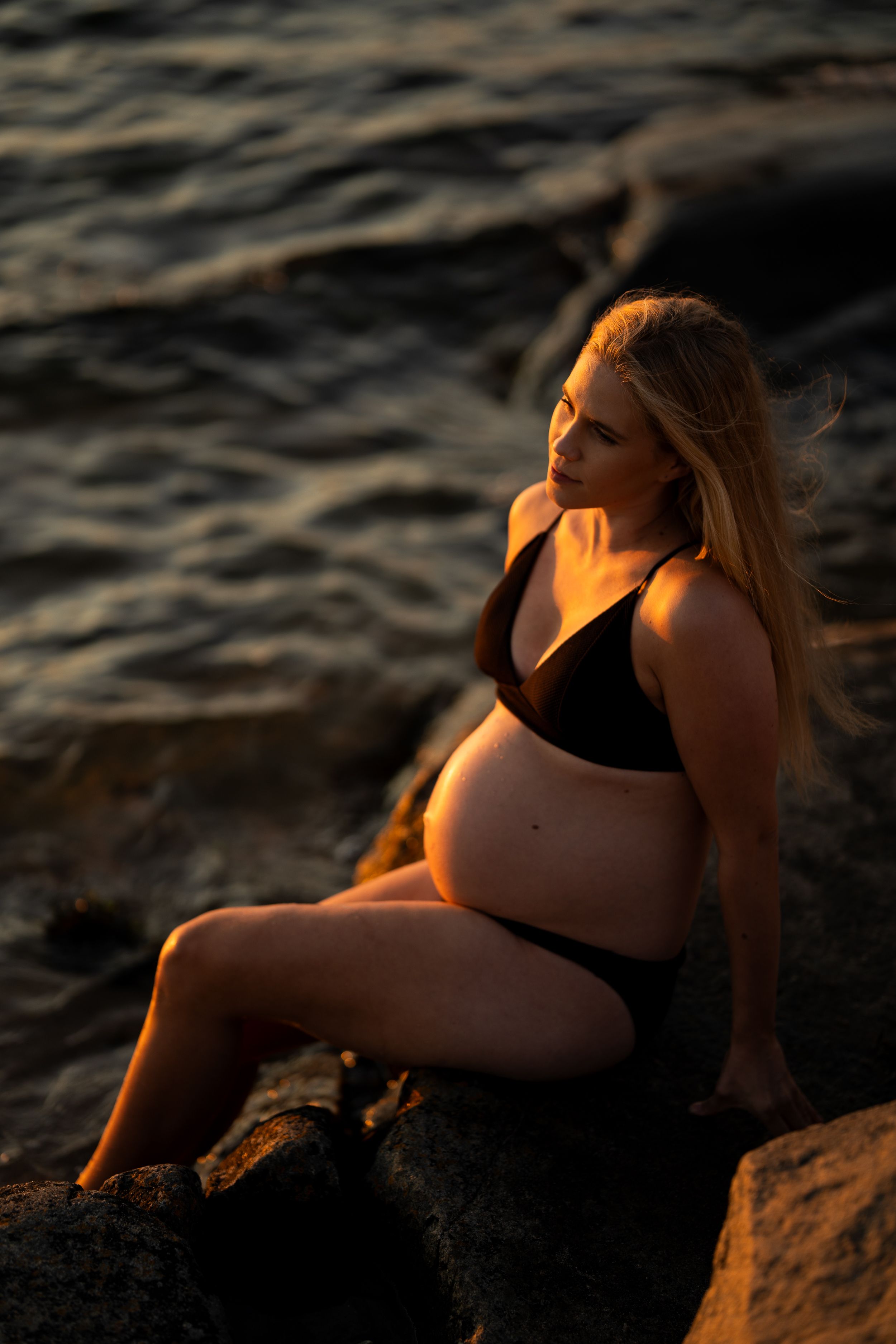 En gravid kvinna i svart bikini sitter på klippor vid vattnet och tittar mot vattnet.