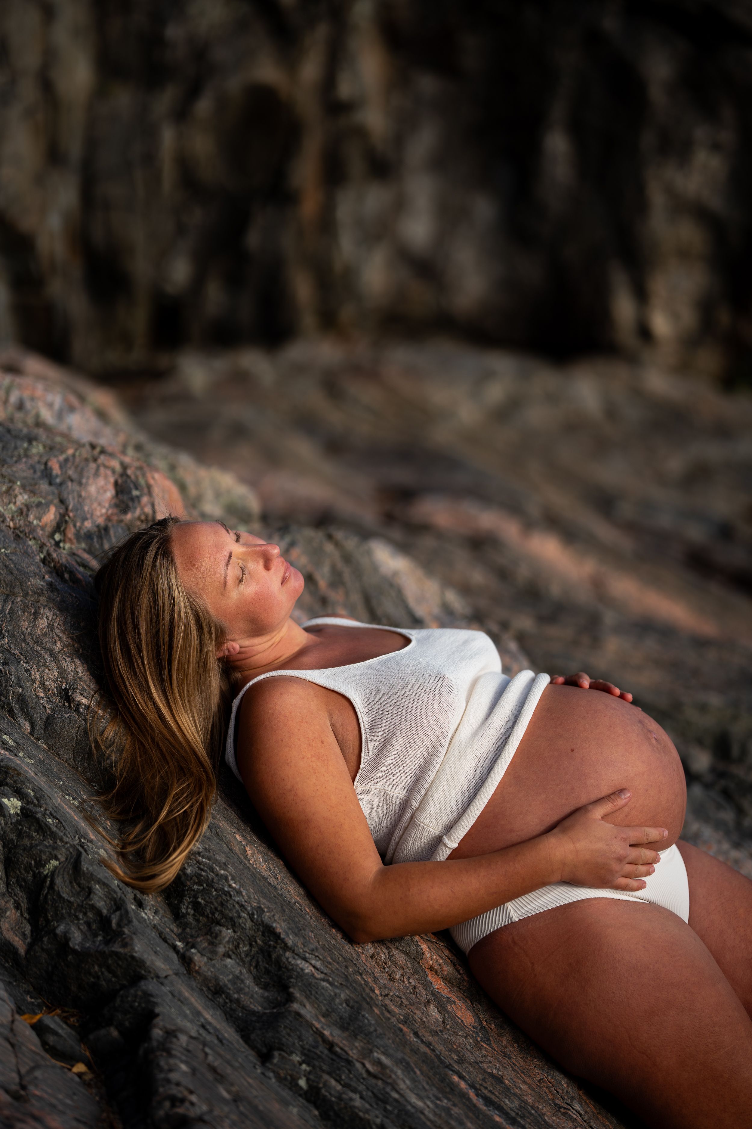 En gravid kvinna i vit topp och vita trosor ligger på en klippa och håller på magen.