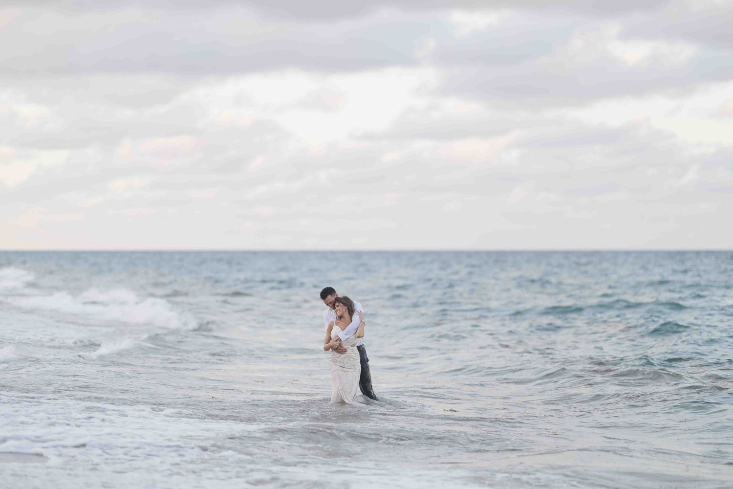 A romantic couple embraces in waist-deep ocean water against a cloudy sky.