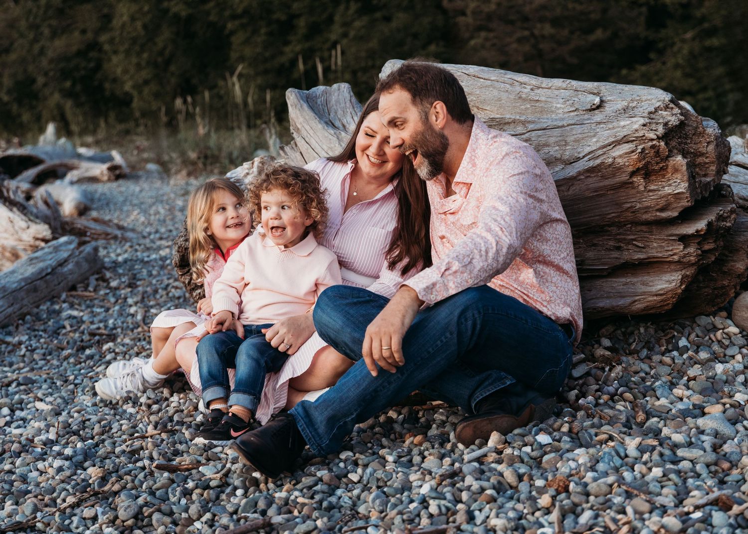 Family sitting together on driftwood at a rocky beach during sunset.