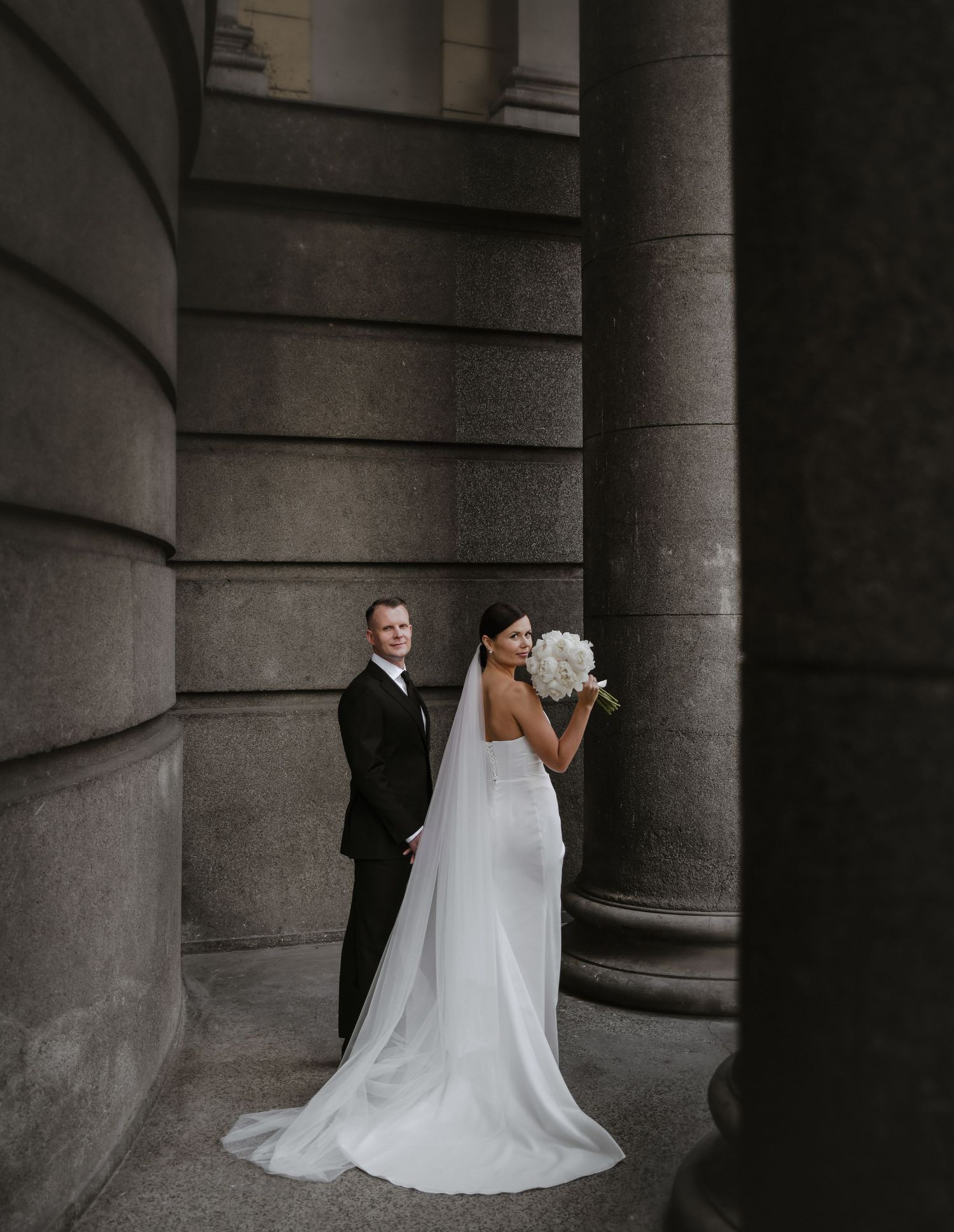 A couple poses elegantly against stone columns during their wedding photoshoot.