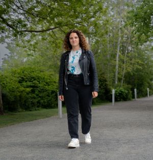 Person in leather jacket and jeans walking on path with green trees in background.