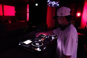 DJ performs at a nightclub using turntables and mixing equipment in a dimly lit room with red ambient lighting.