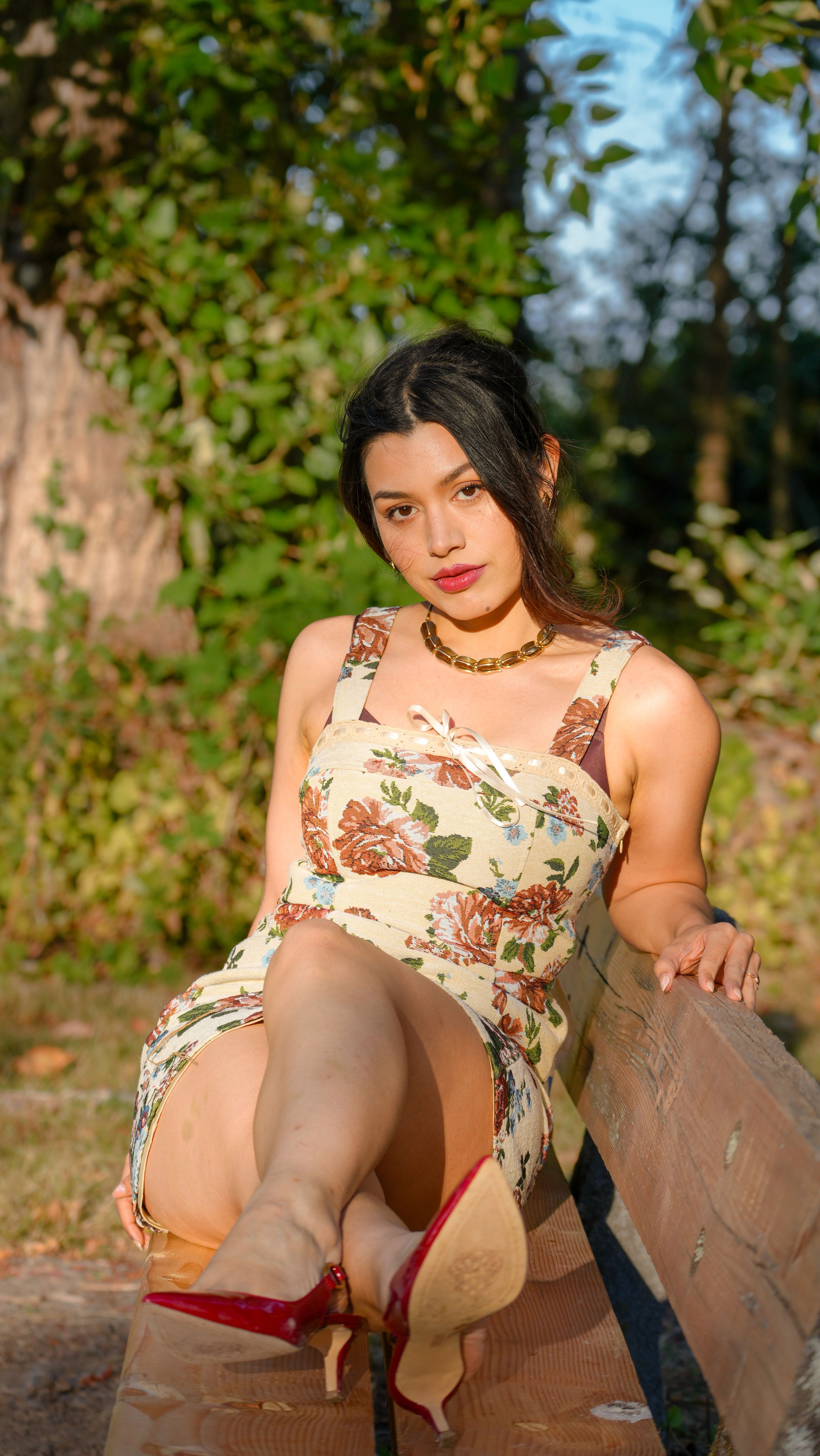 A person in a floral sundress sits on a bench wearing red heels, with sunlit foliage in the background.