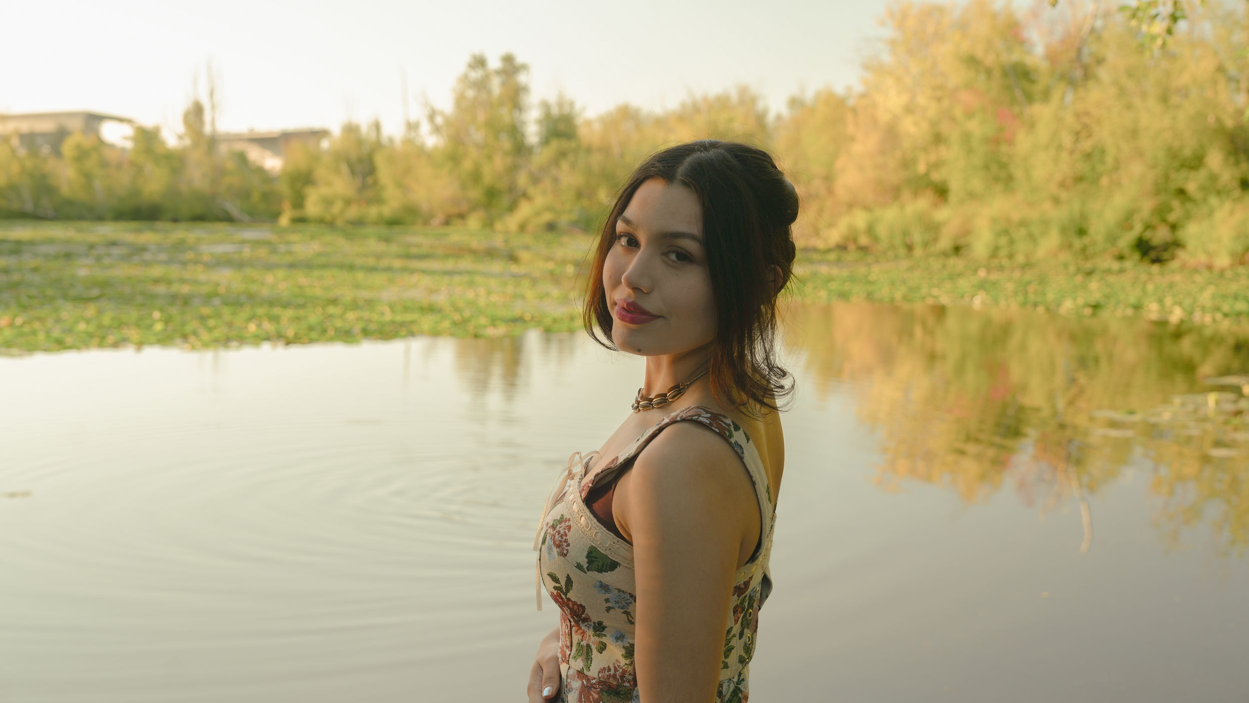 A person in a floral dress stands gracefully on wooden decking beside a serene lake at golden hour.