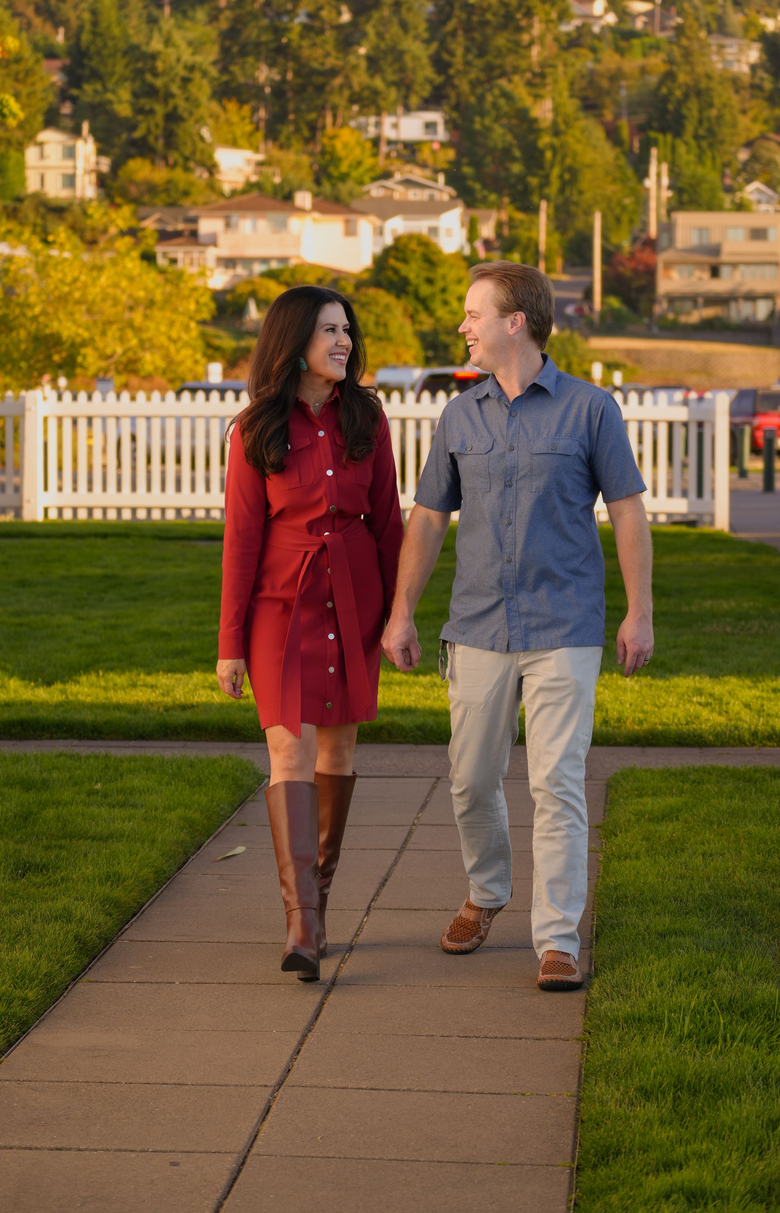 A couple walks hand-in-hand on a sunny pathway, smiling at each other, with green lawns and a white fence behind them.