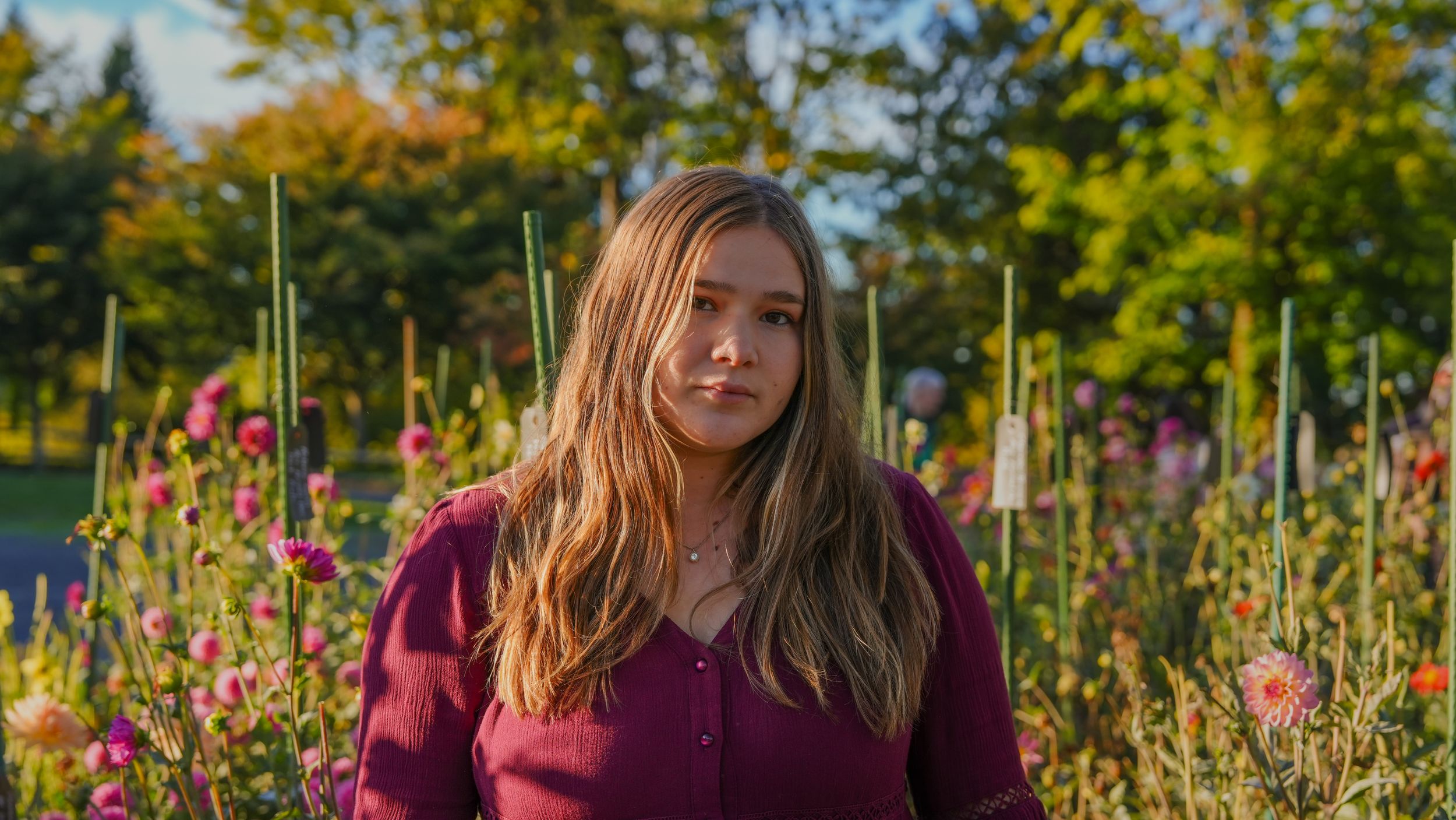 Person in burgundy cardigan stands in a garden with pink hollyhock flowers and autumn foliage in background.