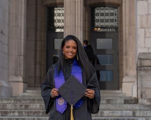 Graduate in black cap and gown with purple stole proudly displays diploma in front of academic building.