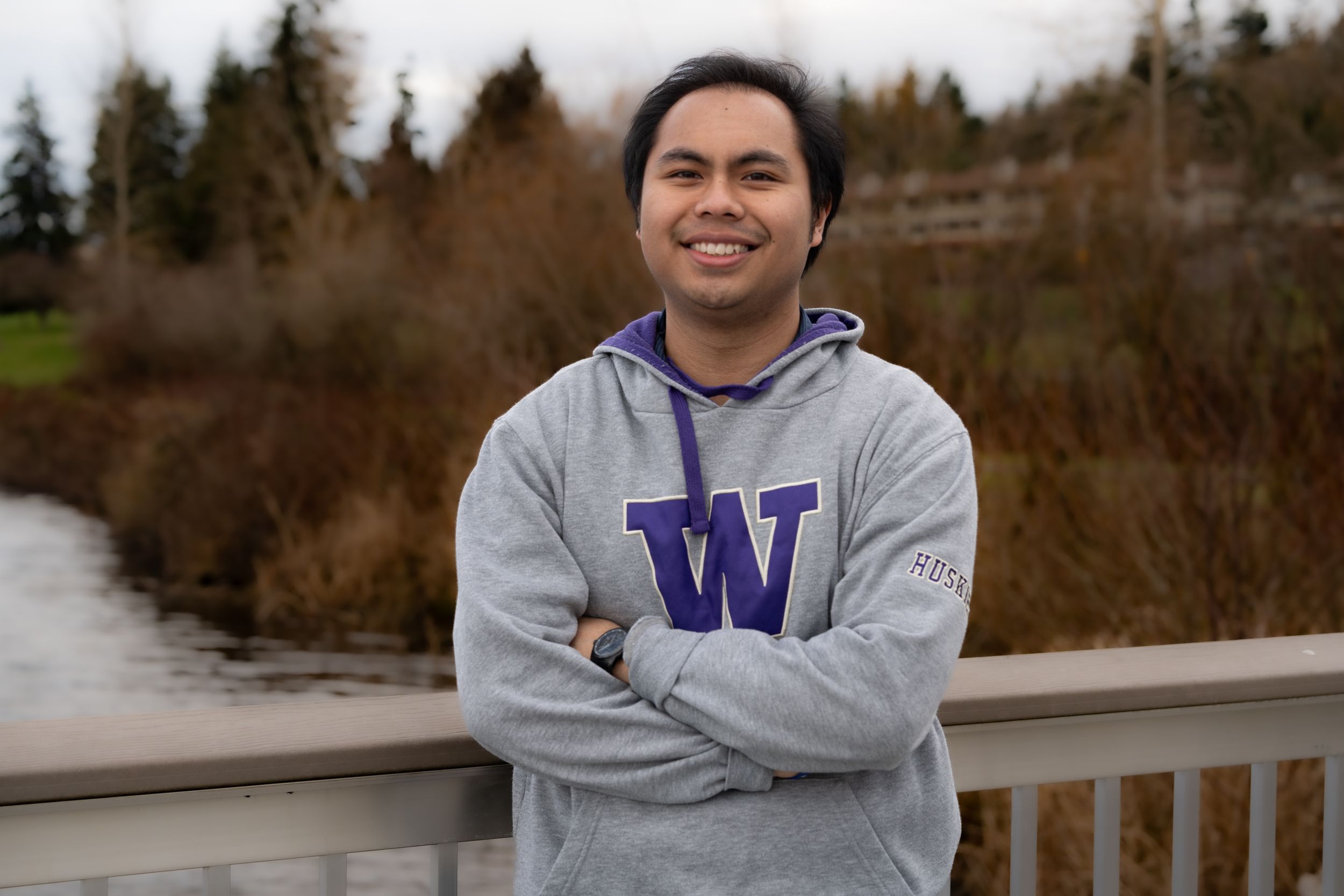 Person in a grey University of Washington sweatshirt stands on a bridge overlooking nature.