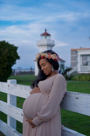 Expectant mother in flowing dress with flower crown poses by white fence near lighthouse.