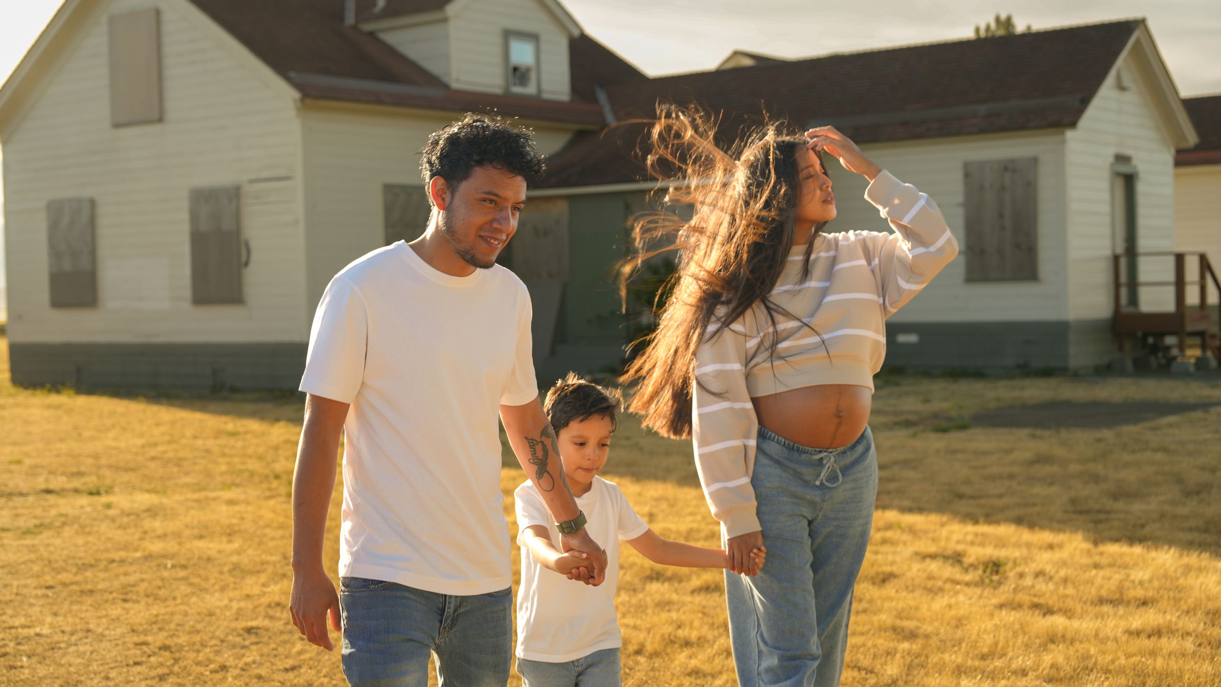 Family of three walking outdoors in golden sunlight near a white farmhouse on a sunny day.