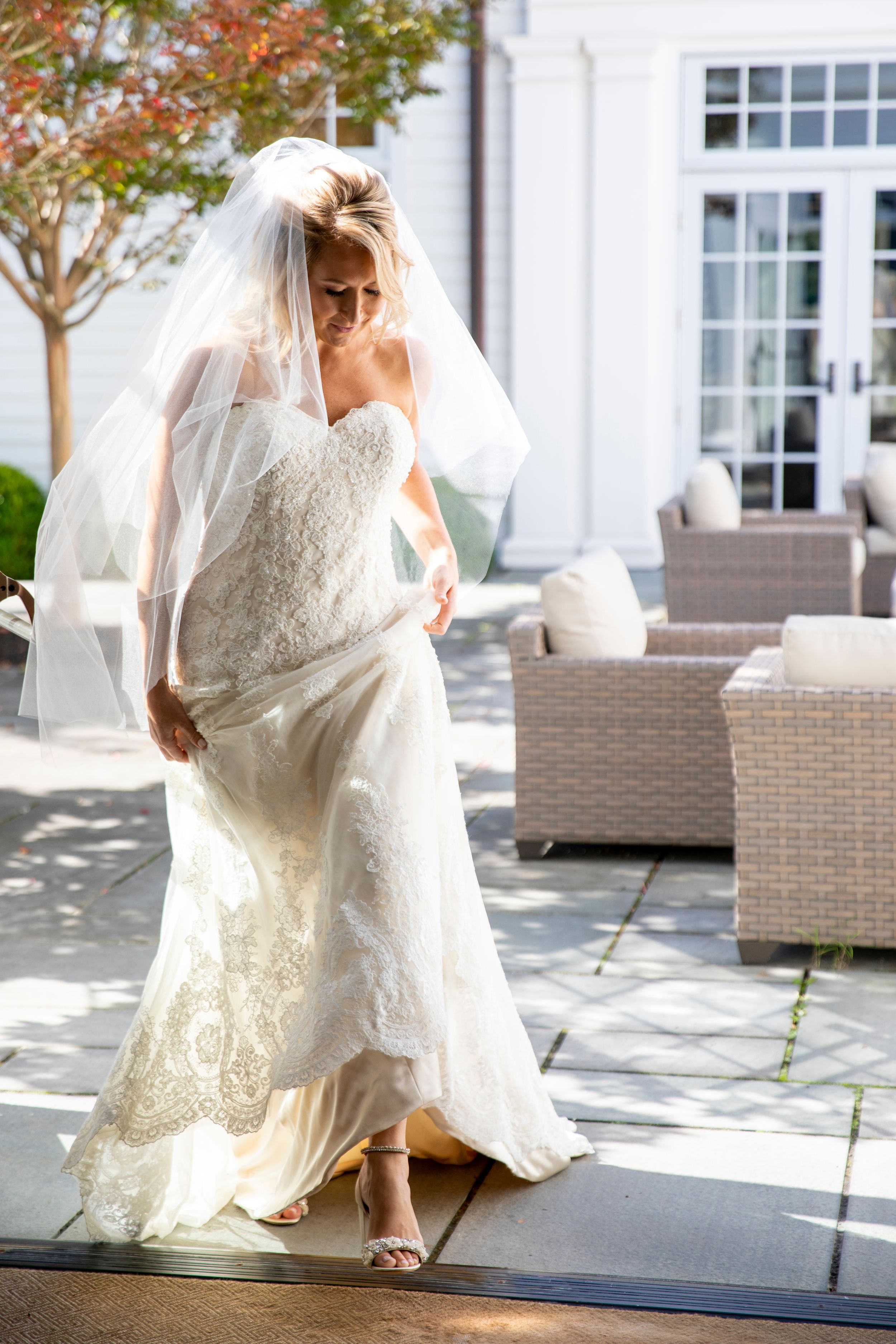 Elegant bride in lace wedding dress walking on outdoor patio with flowing veil.
