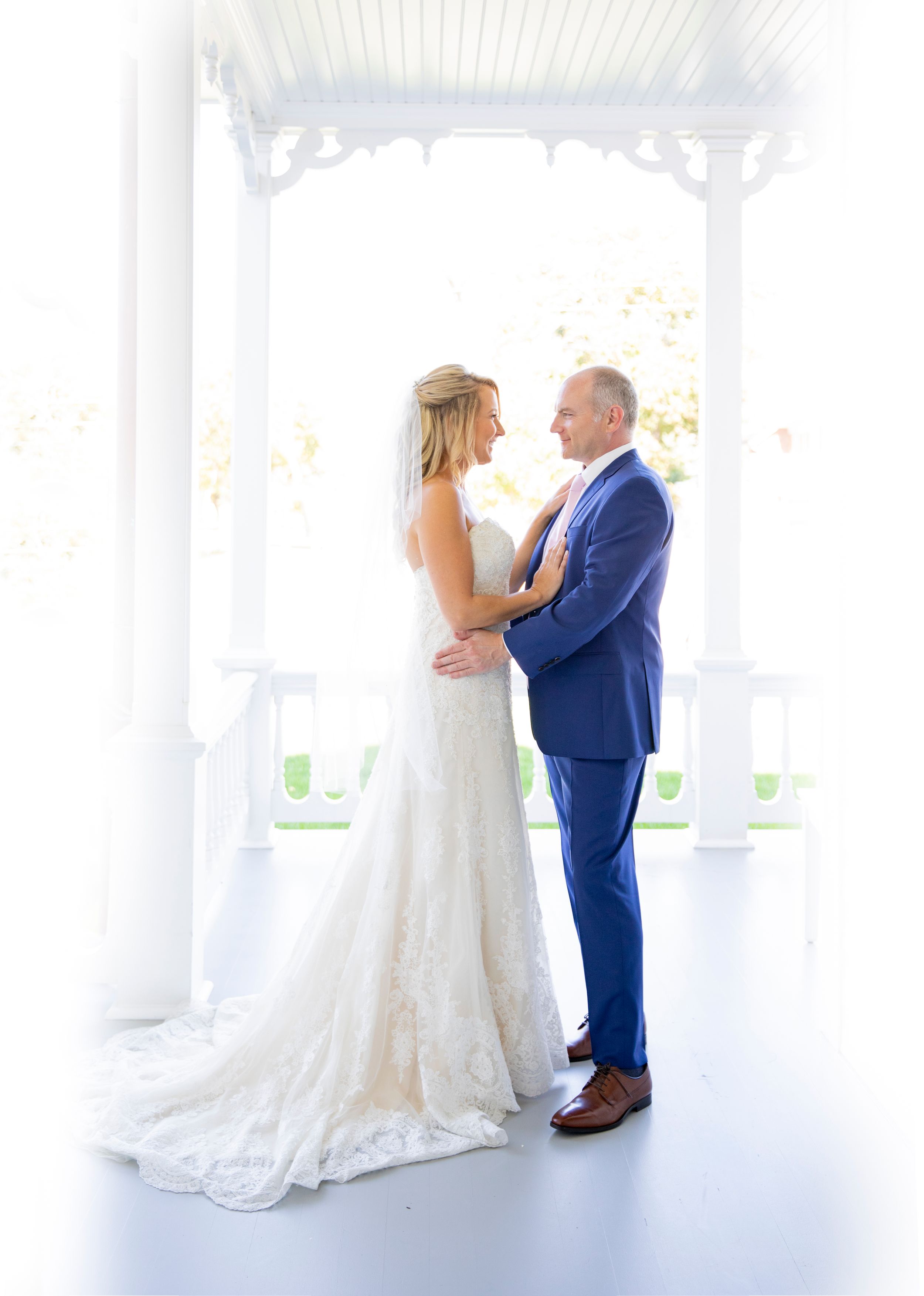 Bride in lace gown and groom in blue suit share romantic moment on white porch.