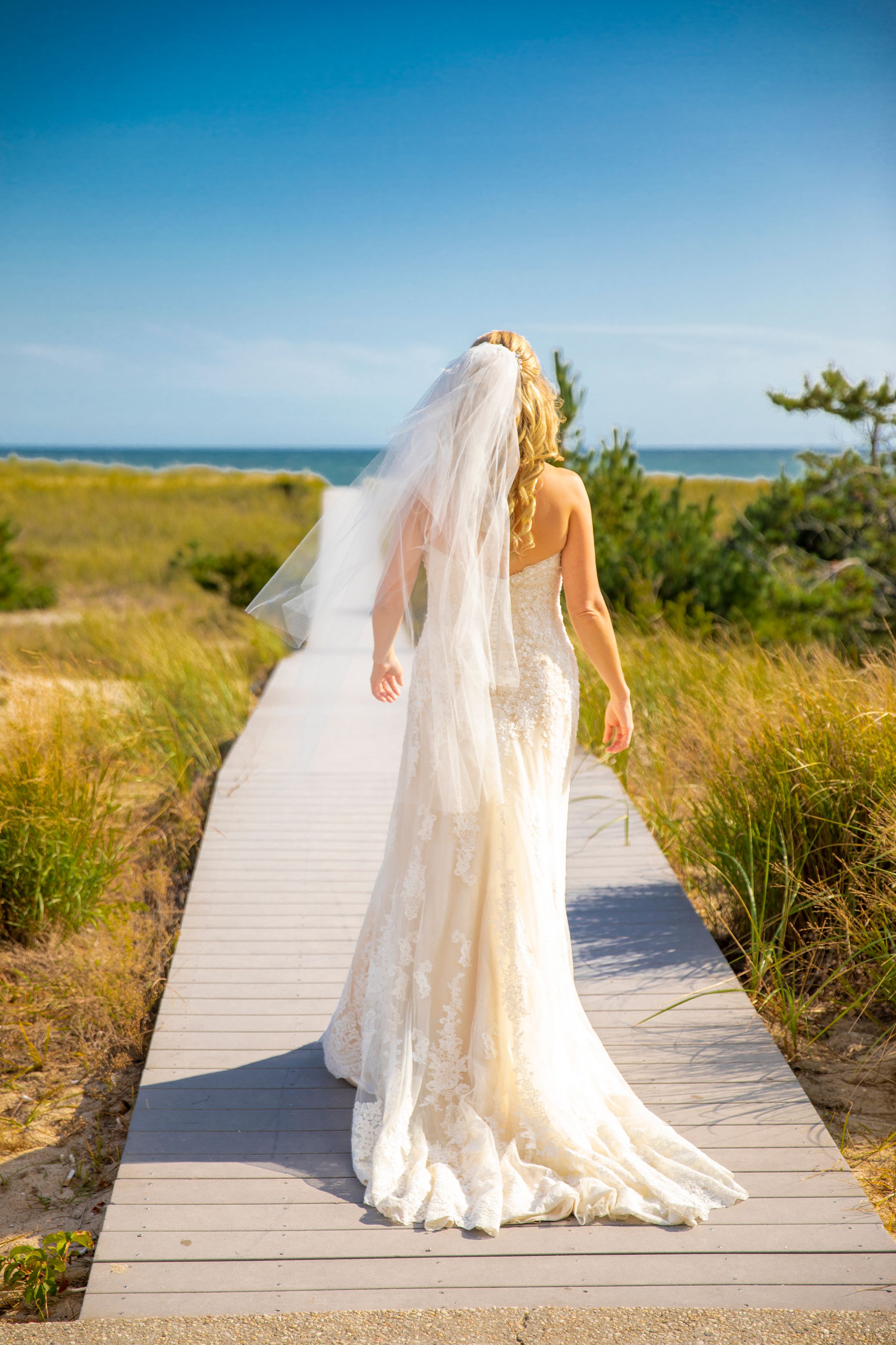 Bride in flowing lace gown walking on coastal boardwalk with ocean view.